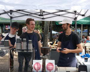 Tres jóvenes baristas con delantales sirven café y preparan bebidas en una carpa al aire libre en un puesto de mercado.