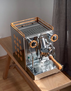 Máquina de Espresso de acero inoxidable con detalles de madera colocada sobre una mesa de madera cerca de una ventana con cortina gris.