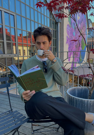 Hombre sentado en un balcón con taza de café en la mano leyendo un libro sobre café en un día despejado.