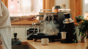 Persona usando máquina de Espresso en una cocina luminosa con vapor de la preparación y accesorios de café al fondo.