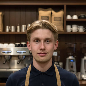 Joven con uniforme de Barista frente a máquinas de Espresso y estantes con granos de café y tazas en una cafetería.