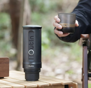 Cafetera espresso portátil en tono gris oscuro para uso exterior con vaso de café en mano, de la categoría de cafeteras portátiles.