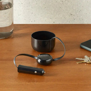 Cargador de coche para taza eléctrica Ember colocado sobre mesa de madera con adaptador para coche y detalles reflectantes.